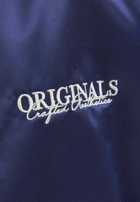 Mornarska tkanina s bijelom vezom i natpisom "ORIGINALS Crafted Aesthetics." Glatka tekstura s blagim sjajem.