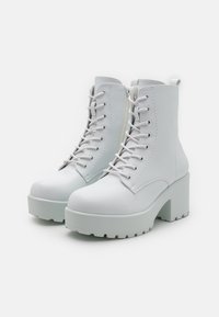 Hvite høye hæler laget av syntetisk materiale. Funksjoner inkluderer rund tå, snøring foran, glidelås på siden og tykk plattformsåle.