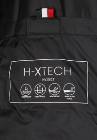 Panneau en tissu noir avec étiquette indiquant les caractéristiques de protection H-XTECH : imperméable, respirant, coupe-vent et icônes des coutures principales étanchées.