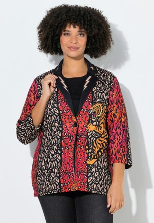 Kvinde med krøllet hår iført en flerfarvet jakke med dyreprint med en tiger-grafik og lynformet kravedetalje.