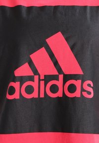 Svart tygtröja med kort ärm och en iögonfallande röd Adidas-logotyp som har tre diagonala ränder ovanför varumärkesnamnet i matchande röd färg.
