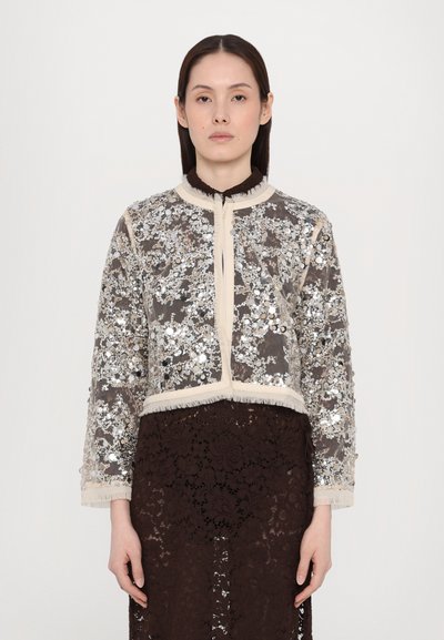 Jachetă scurtă din material transparent, embellizată cu paiete argintii și modele florale, cu o margine bej și guler franjurat. Fustă din dantelă maro dedesubt.