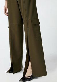 Maslinasto zelene široke hlače s cargo džepovima, sa bočnim prorezima i glatkom teksturom. Uparene s crnim lakiranim cipelama s oštrom točkom.