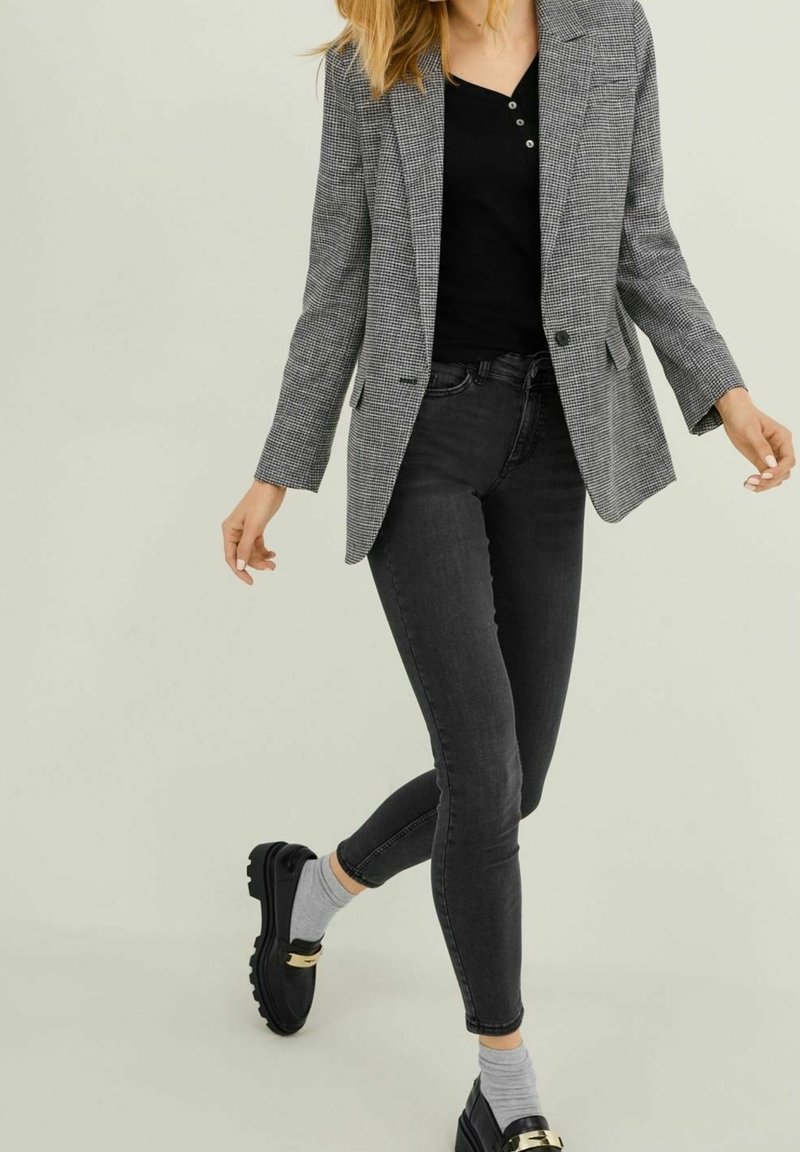 Blazer grigio a quadri con rever a incavo sopra una maglia nera, abbinato a jeans scuri e mocassini neri con dettagli dorati. Texture lisce in tutto l'insieme.