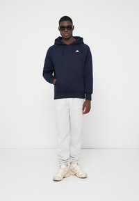 Άνδρας με ναυτικό φούτερ, ανοιχτό γκρι φόρμες, μπεζ αθλητικά παπούτσια και μαύρα γυαλιά ηλίου, που στέκεται μπροστά σε ένα λευκό φόντο.