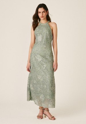 Joven mujer con un vestido maxi sin mangas, de cuello alto, con un estampado verde claro y sandalias de tacón abiertas delante de un fondo liso.