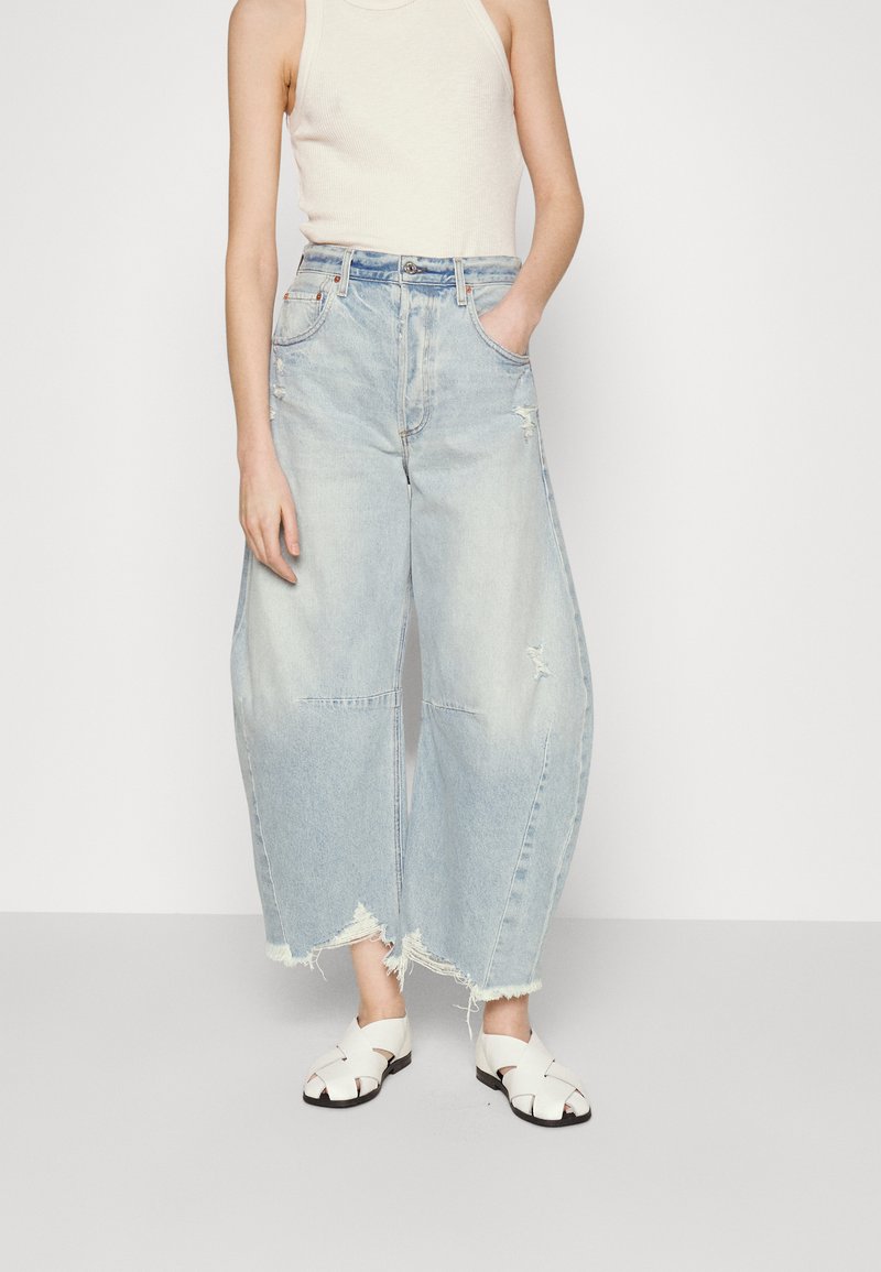 Jeans in denim chiaro a gamba larga con orli sfrangiati e dettagli strappati, abbinati a sandali bianchi con cinturini e un top beige aderente.