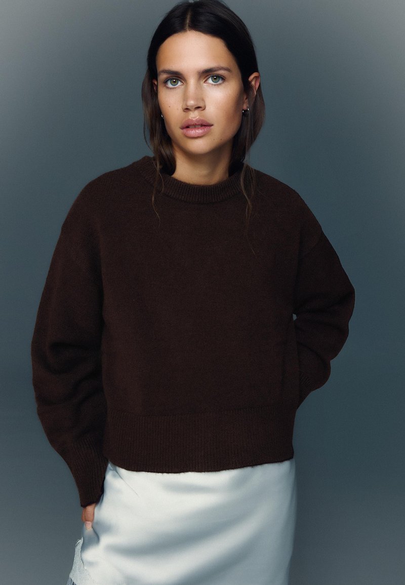 Pulover maro supradimensionat cu decolteu rotund și manșete cu nervuri, combinat cu o fustă ușoară din satin, pe un fundal gri.