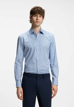 Jonge man staat en kijkt vooruit, draagt een blauw bloemenoverhemd met lange mouwen en een donkere broek tegen een effen lichte achtergrond.