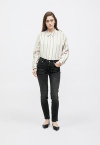 Camisa de botão branca com riscas em tons pastel, jeans pretos justos e sapatos planos pretos. A camisa apresenta uma gola clássica e mangas com punhos.