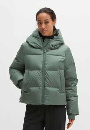 Femme portant une doudoune vert sauge avec une grande capuche, un pantalon noir et un haut noir, debout devant un fond blanc.