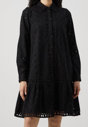 Vrouw draagt een zwarte ooglet-jurk met lange mouwen, een kraag, knopen aan de voorkant en uitgesneden kanten details aan de zoom en taille.