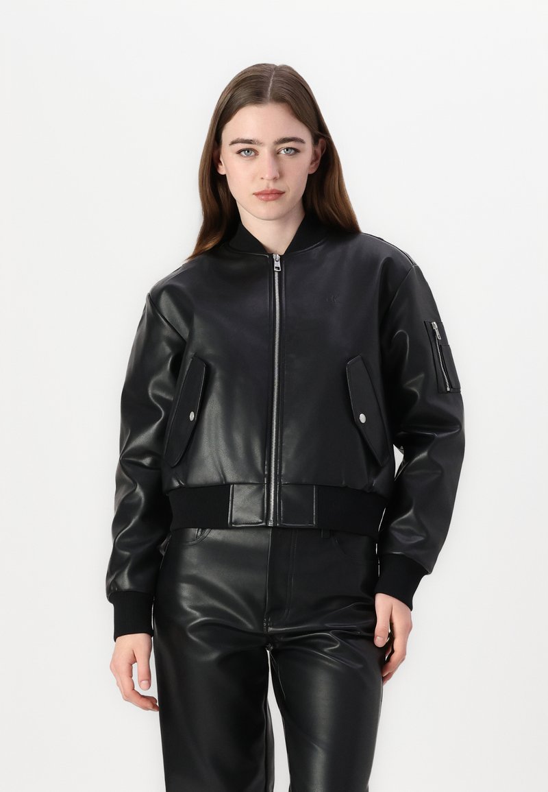 Giacca bomber nera in ecopelle con zip frontale, polsini a costine e due tasche frontali con chiusura a scatto. Texture liscia, design corto.