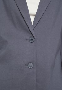 Szara marynarka wykonana z gładkiej tkaniny, posiadająca klapy z wycięciem oraz dwa niebieskie guziki do zapinania. Widoczny jest subtelny detal szwu.