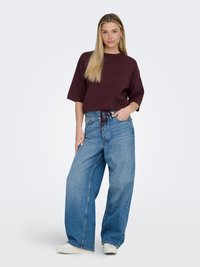 Jeune femme portant un jean bleu ample, un haut à manches courtes marron foncé et des baskets blanches, debout avec une main dans la poche.