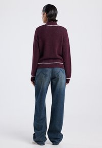 Persoon die weggekeerd staat, gekleed in een kastanjebruine trui met witte accenten en ruime blauwe jeans, tegen een effen achtergrond.