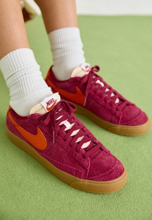 Personne portant des chaussettes blanches et des baskets Nike bordeaux avec un swoosh orange et des semelles en gomme sur un sol recouvert de moquette verte.