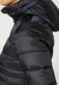 Nærbillede af en person iført en sort quiltet pufferjakke med høj krave og et rundt logo-patch på overarmen.