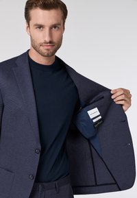 Giacca da abito texture blu navy con due bottoni, aperta per rivelare una tasca interna e un’etichetta di tessuto; abbinata a una camicia blu fitted.