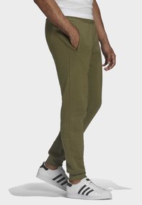 Olivgröna sweatpants med sidfickor, avsmalnande passform och mjuk textur, kombinerade med vita sneakers med svarta ränder och texturerad sula.