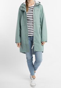 Ljusgrön vattentät jacka med huva, dragkedjestängning och sidofickor. Bärs över en svart och vit randig skjorta och blå jeans.