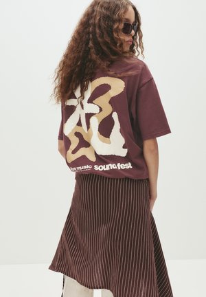 Bordeauxrotes Oversized-T-Shirt mit abstrakten cremefarbenen und beigen Mustern auf dem Rücken, kombiniert mit einem gestreiften bordeauxroten Rock. Text ist auf dem Shirt vorhanden.
