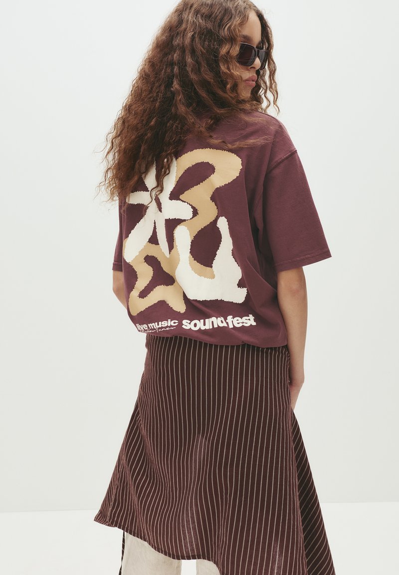 Bordeaux oversized t-shirt med abstrakte mønstre i creme og beige på ryggen, parret med en stribet bordeauxnederdel. Der er tekst på t-shirten.