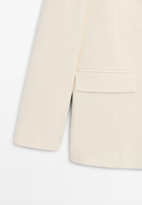 Nærbillede af ærmet på en beige uldblazer og en klaplomme på en ren hvid baggrund, der fremhæver stoffets tekstur og skrædderdetaljer.