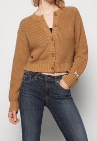 Cárdigan de punto marrón con un diseño corto, bordes acanalados y cierre de botones. Combinado con unos jeans de mezclilla clásicos en azul oscuro.