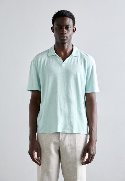 Polo de color azul claro con mangas cortas y una tela suave y texturizada. Presenta un cuello clásico y un corte holgado, combinado con pantalones beige claros.
