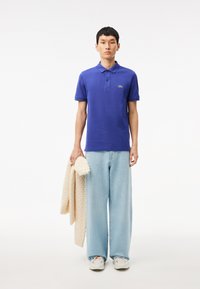 Camisa polo morada con cuello clásico, combinada con unos vaqueros de pata ancha de color azul claro. El modelo sostiene un suéter de punto color crema. Los zapatos son de color gris claro.