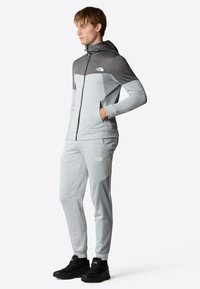 Tuta sportiva grigio chiaro realizzata in tessuto testurizzato; presenta una giacca con cappuccio e dettagli neri e pantaloni affusolati abbinati. Calzature nere.