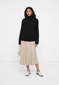 Črn rebrast pulover s turtleneck ovratnikom, bež midi krilo, bele slip-on čevlje, drži kremasto torbico, na belem ozadju.