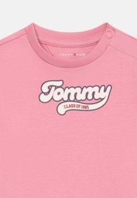 T-shirt di cotone rosa con un grande logo "Tommy" in bianco con contorno nero e "Class of 1985" in rosso. Collo rotondo con chiusura a pressione.