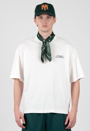 Junger Mann trägt ein weißes oversized T-Shirt mit Text, dunkelgrünes Bandana um den Hals gebunden, passende dunkelgrüne Baseballkappe und grüne Shorts.