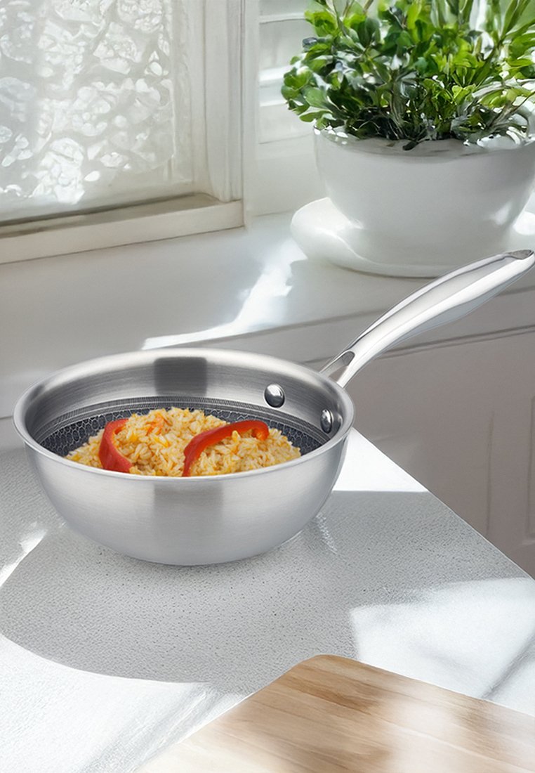 Patelnia ze stali nierdzewnej z polerowaną powierzchnią, zawierająca żółty ryż i plastry czerwonej papryki, na teksturowanym blacie.