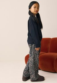 Sort sweatshirt, mønstrede bredbenede leopardprint bukser i grå og taupe, brune ankelstøvler, stående ved siden af en rustfarvet sofa.