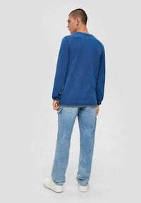Blaues strukturiertes Langarmhemd mit seitlichen Knöpfen, kombiniert mit hellblauen Jeans und weißen Sneakers. Von hinten betrachtet.