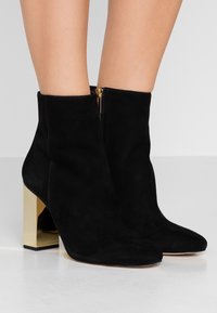 Botas de tobillo de ante negro con puntera afilada, detalle de cremallera lateral y un tacón de bloque dorado. Textura suave con un acabado mate.