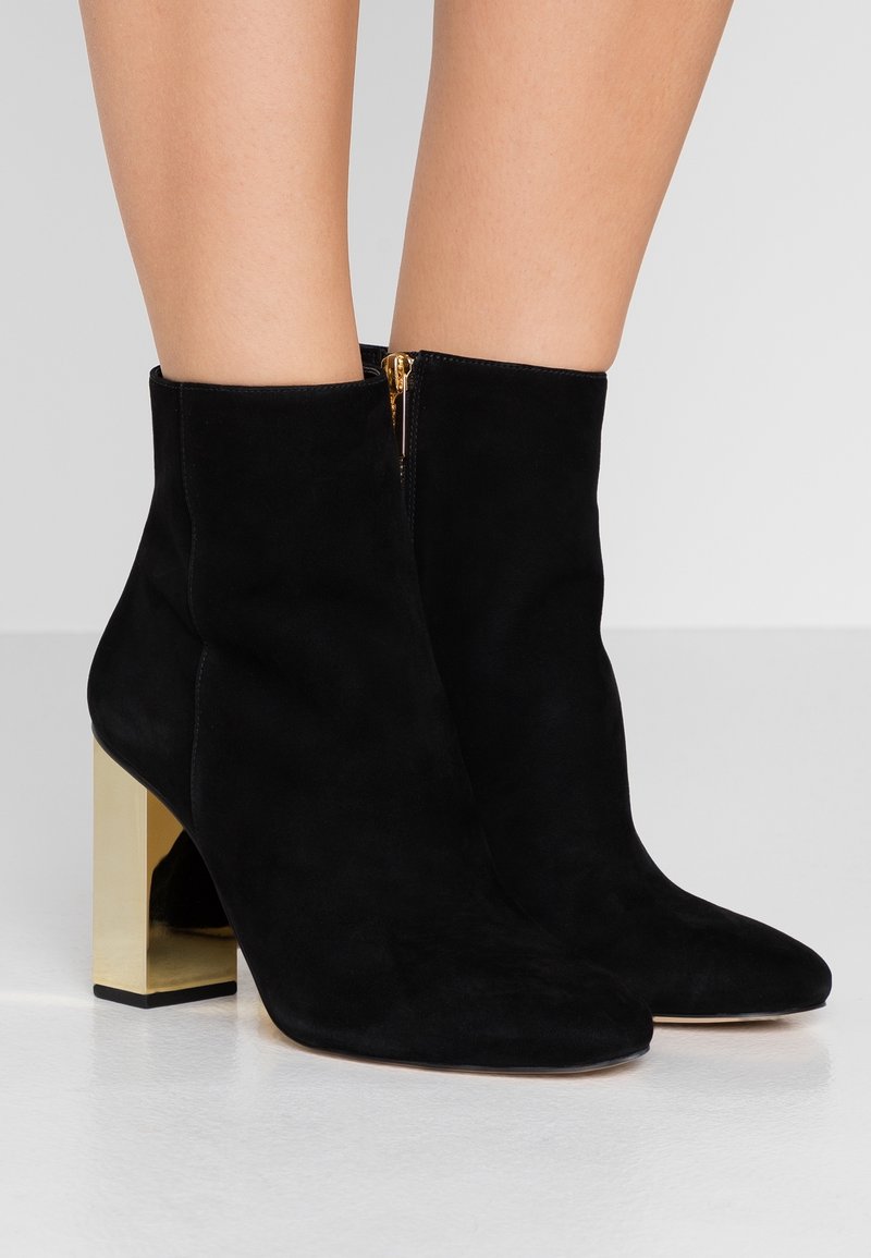 Botas de tobillo de ante negro con puntera afilada, detalle de cremallera lateral y un tacón de bloque dorado. Textura suave con un acabado mate.