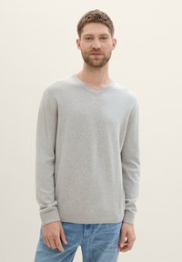 Pull en v gris clair en tissu doux, avec des manches longues et des poignets côtelés, porté avec un jean bleu. Design simple et ajusté.