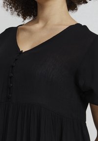 Svart klänning med V-urringning, som har en knäppning med knappar och en texturerad tyg. Designen inkluderar korta ärmar och en utsvängd kjol.