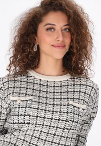 Textúrált pulóver krém és fekete színben, rácsos mintával. Két mellzsebbe varrt díszgombokkal és kerek nyakkivágással rendelkezik.