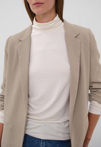 Femme portant un blazer beige sur un pull à col roulé blanc et un jean bleu, montrée des épaules aux hanches sur fond uni.