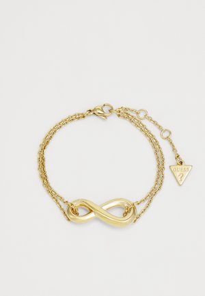 Χρυσή επιχρυσωμένη βραχιόλι που διαθέτει σύμβολο του άπειρου, διπλό σχέδιο αλυσίδας και τριγωνικό γουράκι με το "GUESS" τυπωμένο πάνω του.