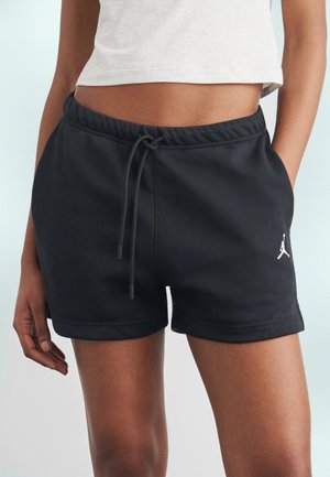 Frau trägt schwarze Sportshorts mit Kordelzug und weißem Jordan-Logo, kombiniert mit einem hellgrauen bauchfreien Oberteil, die Hände in den Taschen.