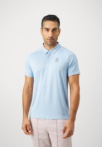Mann som står mot hvit bakgrunn iført lyseblå kortermet polo shirt og lyserosa bukser, vender seg fremover med nøytral uttrykk.