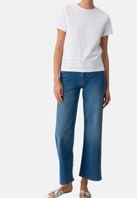 T-shirt blanc à manches courtes avec des rayures horizontales texturées, assorti à un jean en denim bleu à coupe décontractée et des sandales décoratives en argent.