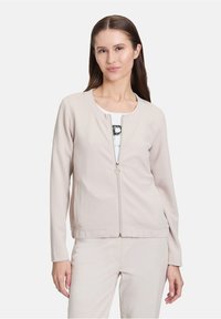 Veste beige clair zippée avec un tissu texturé et respirant. Elle possède un col rond, des manches longues et des poches sur les côtés.