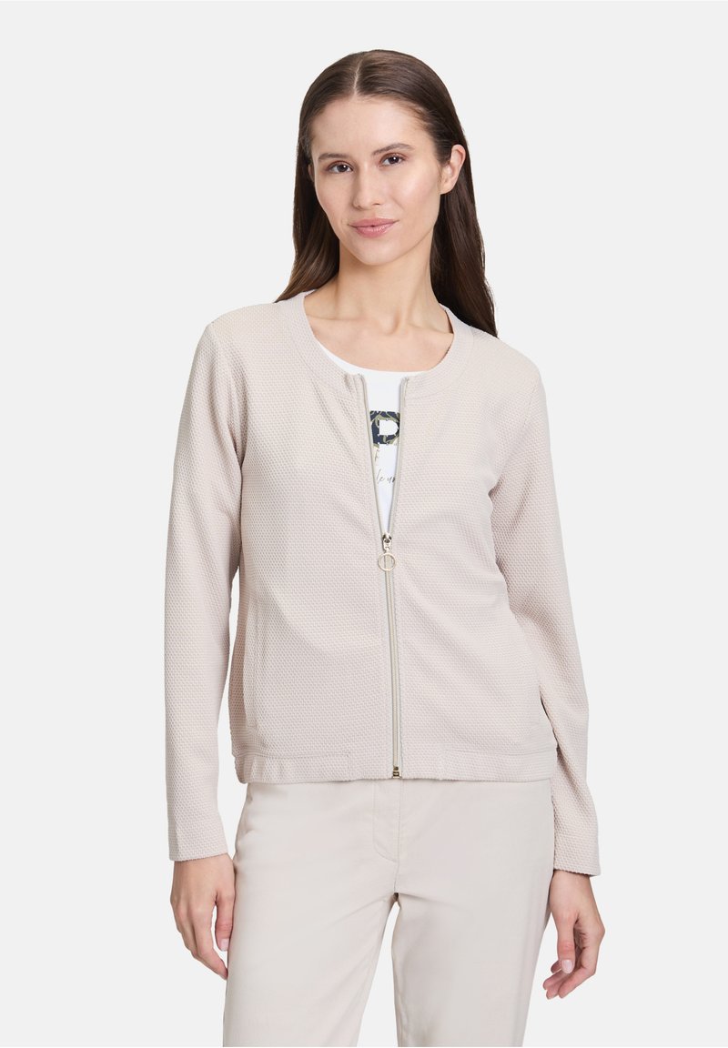 Veste beige clair zippée avec un tissu texturé et respirant. Elle possède un col rond, des manches longues et des poches sur les côtés.
